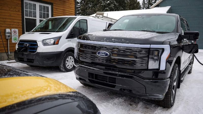 A black Ford F-150 truck and a white Ford Transit van parked in a snowy area near a wooden building, with charging stations visible.