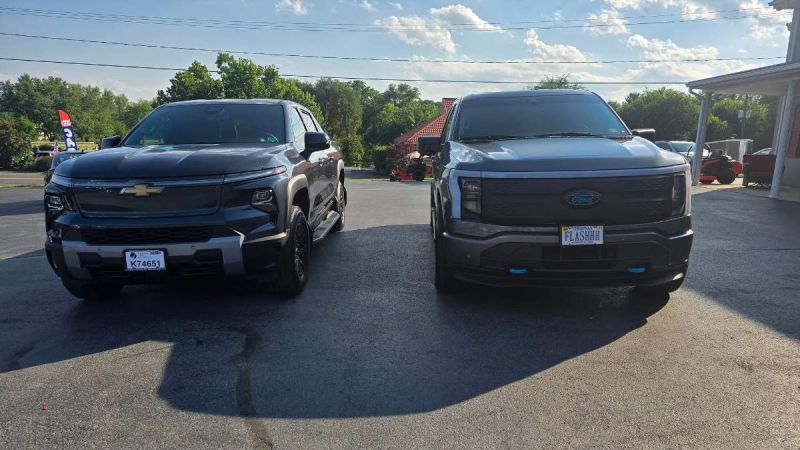 Black Chevy Silverado and Ford F-150 Lightning electric trucks parked side-by-side on asphalt lot with trees and blue sky background