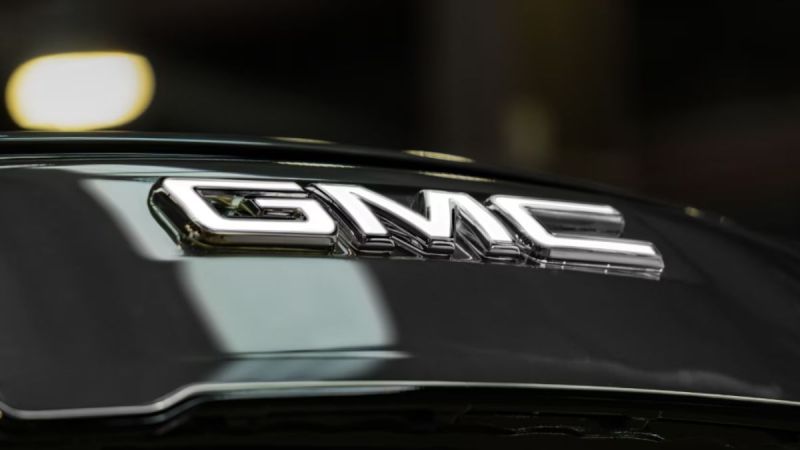 Close-up of a shiny chrome GMC logo on a Sierra EV surface, conveying a sense of luxury and modernity with soft, blurred lighting in the background.