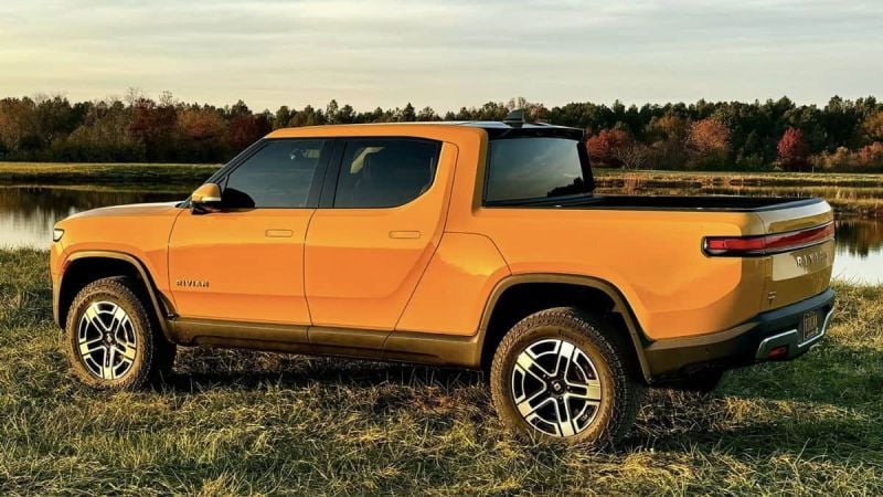 Orange Rivian R1S electric SUV shown from the rear three-quarter view, parked on grass near a lake with autumn trees in the background during sunset.