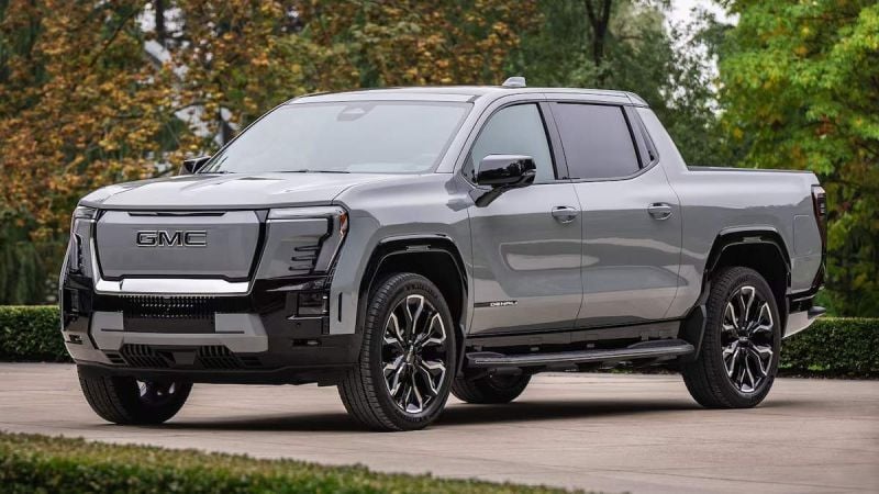 A gray 2025 GMC Sierra EV pickup truck parked outdoors, showcasing its modern design and bold front grille against a blurred natural backdrop.