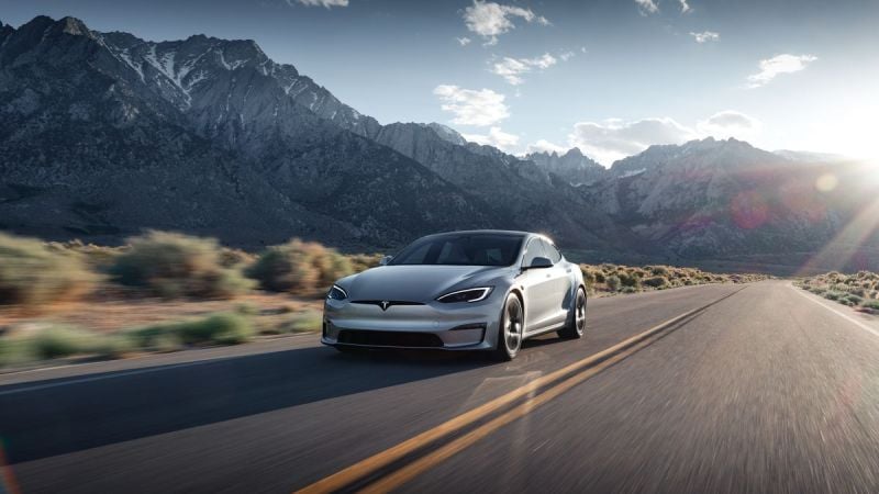 A gray 2025 Tesla Model S drives along a winding road with majestic mountains in the background under a bright sky.