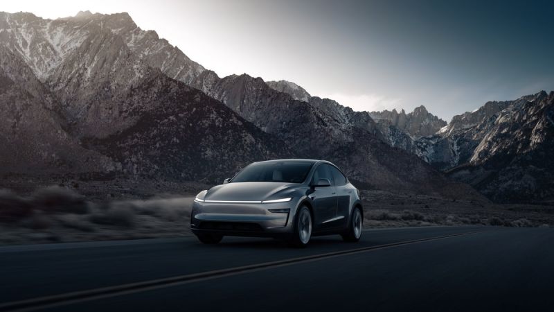 A sleek electric car drives on a deserted road with dramatic mountain peaks in the background, illuminated by soft sunset light.