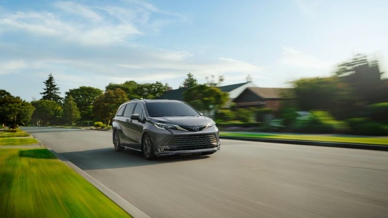 A dark gray 2024 Toyota Sienna minivan shown from the front three-quarter angle, driving on a suburban road with motion blur effect and green landscaping in the background.