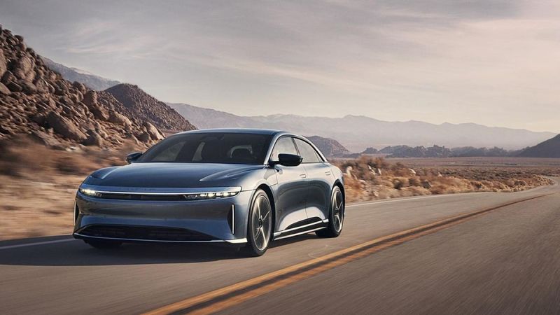 A Gray 2025 Lucid Air drives along a winding road, surrounded by rugged mountains and a vast desert landscape under a clear sky.