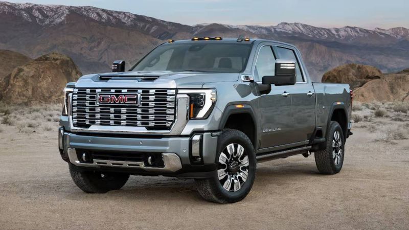 A silver GMC truck parked in a desert landscape, showcasing its bold grille and rugged design against a mountainous backdrop.