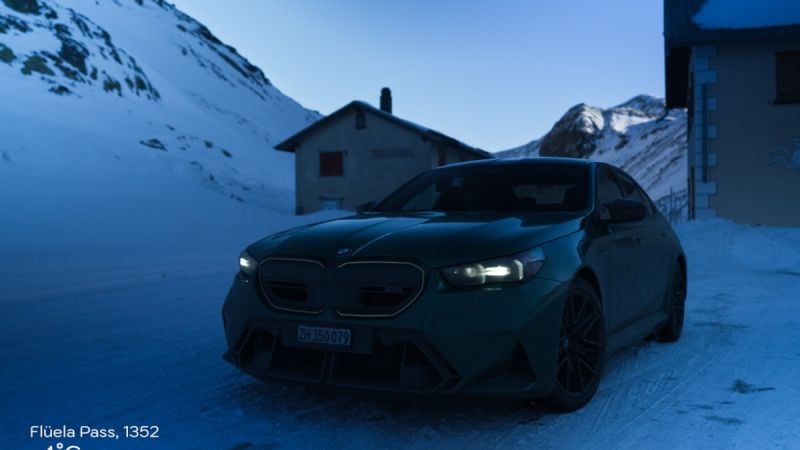 A Green 2025 BMW M5 parked on a snowy road at Flüela Pass, surrounded by mountains under a twilight sky. The car is slightly covered in dirt.