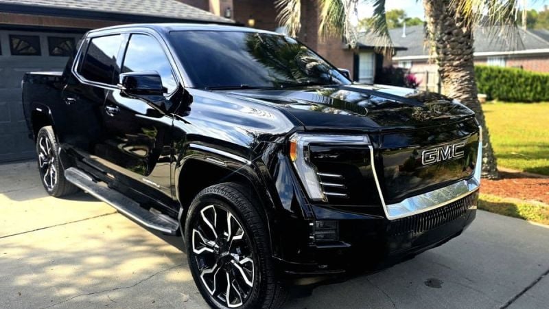 A black GMC Sierra EV shown from a front three-quarter angle parked in a residential driveway, featuring distinctive black and chrome wheels and LED lighting elements.
