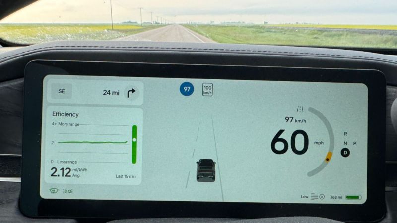 Rivian R1T dashboard showing a digital display. Speed is 60 mph, energy use is 2.12 mi/kWh, with 24 miles to the next turn. Background shows open road with luscious flat-land greenery.