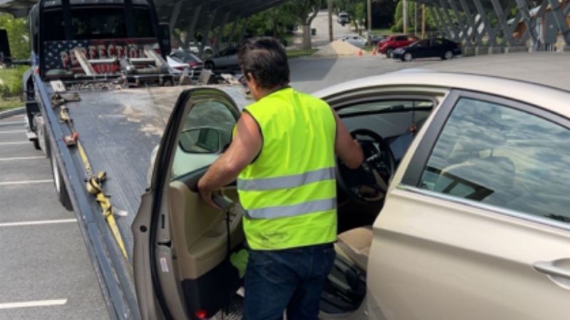 A man in a yellow safety vest prepares to drive a beige Hyundai Sonata onto a tow truck. The scene is in a parking lot with a few parked cars nearby.
