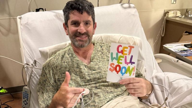 Jack's dad in a hospital bed smiles, holding a colorful "Get Well Soon" card. He gives a thumbs-up with his other hand. The mood is positive and hopeful.