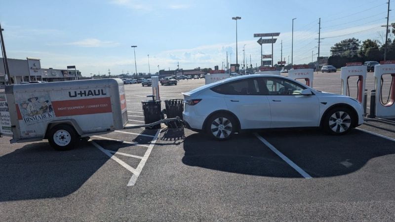 A white Tesla Model Y is parked at a charging station while towing a U-Haul trailer. The scene is in a parking lot under a clear blue sky, conveying a sense of travel.