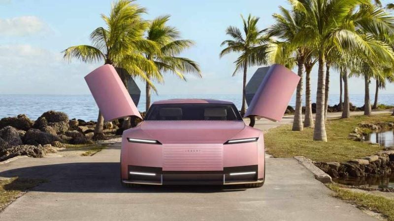 A Pink Jaguar Type 00 with butterfly doors stands on a coastal path, framed by palm trees and rocky shorelines under a clear sky.