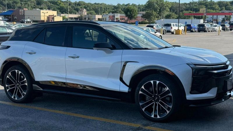 A white Chevrolet Blazer EV is shown from a front three-quarter angle in a parking lot, featuring distinctive black accents and two-tone alloy wheels.