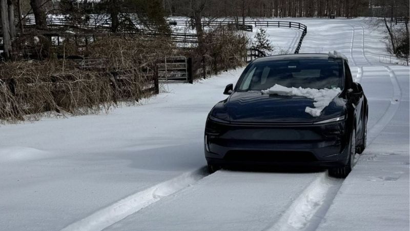 A black Tesla Model Y is parked in a snow-covered driveway, photographed from a three-quarter front angle with winter fencing and trees visible in the background.