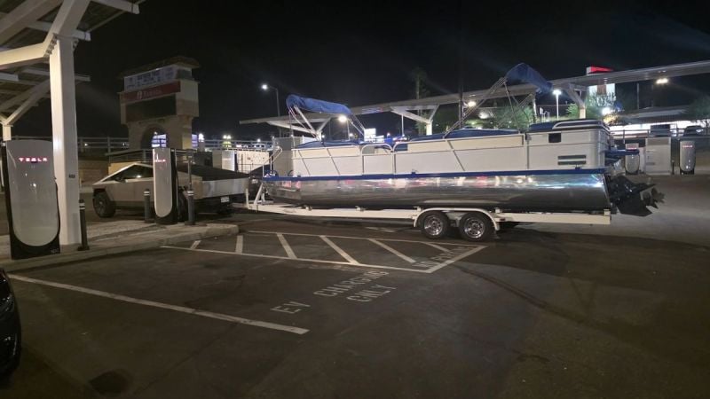 A boat is being towed by a Tesla Cybertruck parked in a charing station at night, the scene is illuminated by overhead lighting.