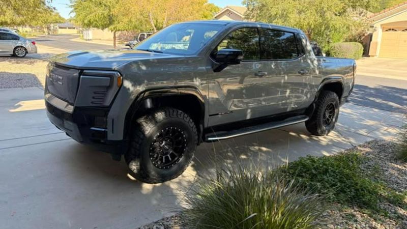 Gray GMC Sierra EV with black off-road wheels, lifted suspension, and custom front bumper parked on residential driveway in suburban setting.