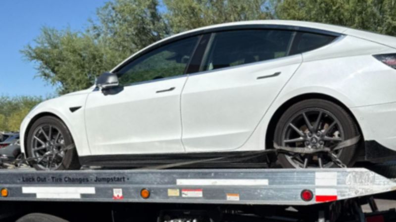 A white Tesla Model 3 is secured on a flatbed tow truck in a sunny outdoor setting with trees in the background, suggesting it needed roadside assistance.