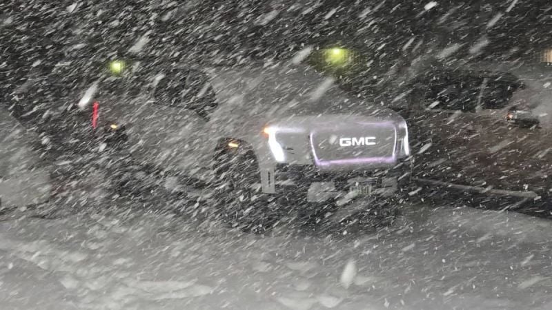 A GMC Sierra EV photographed during heavy snowfall at night, with its illuminated GMC badge visible through the swirling snow and yellow lights glowing in the background.