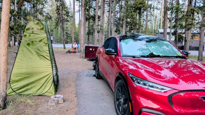 A red Ford Mustang Mach-E is parked in a forest campsite near a green tent. A person sits by a fire pit in the background, surrounded by trees and cabins. The scene feels peaceful and rustic.