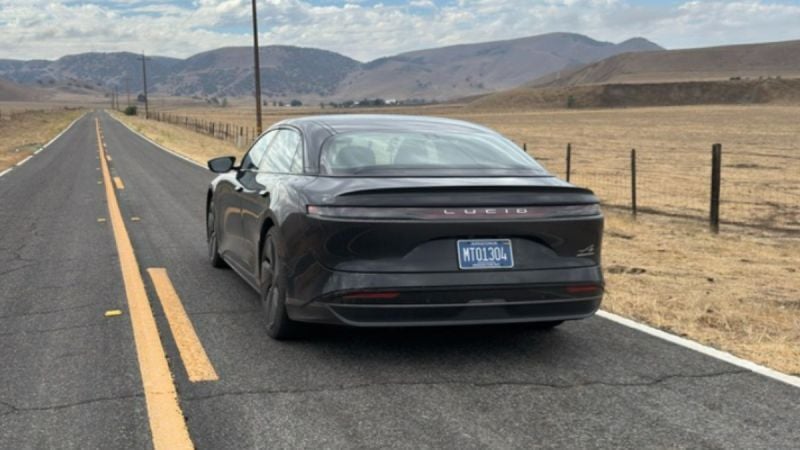 A black Lucid Air drives down an empty rural road flanked by dry fields and distant hills under a partly cloudy sky, conveying a sense of freedom.