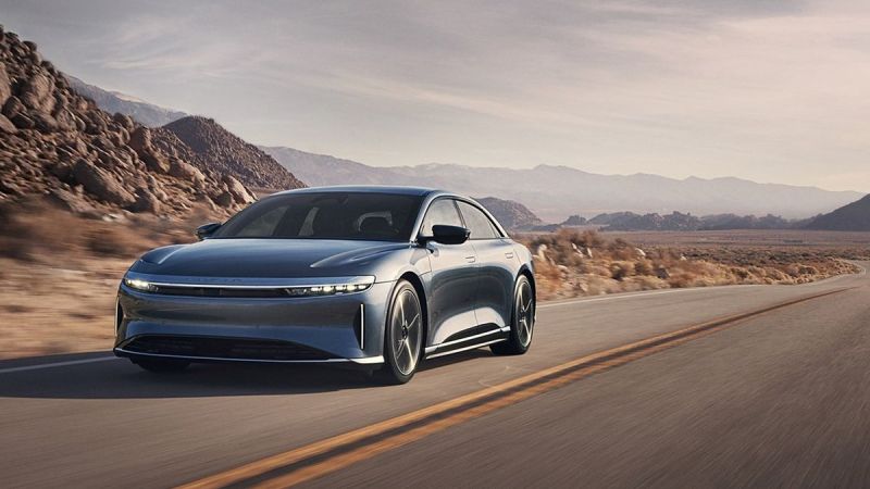 A sleek Lucid Air driving along a winding road with rocky mountains and a hazy sky in the background.