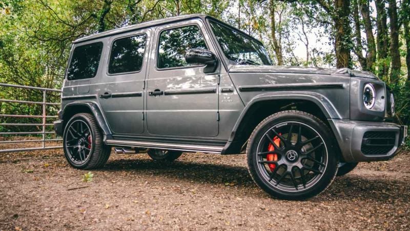 A gray Mercedes-Benz G-Class SUV shown from a side-front quarter view, featuring black aftermarket wheels, red brake calipers, and photographed in a wooded setting.