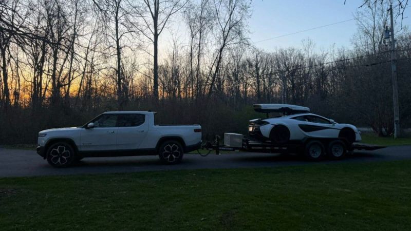 White Rivian R1T towing a trailer with a McLaren at sunset, surrounded by bare winter trees