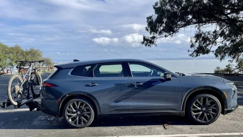 A blue Toyota Crown Signia shown from the side view, featuring black wheels and a bike mounted on a rear rack, parked in an oceanside lot under partly cloudy skies.