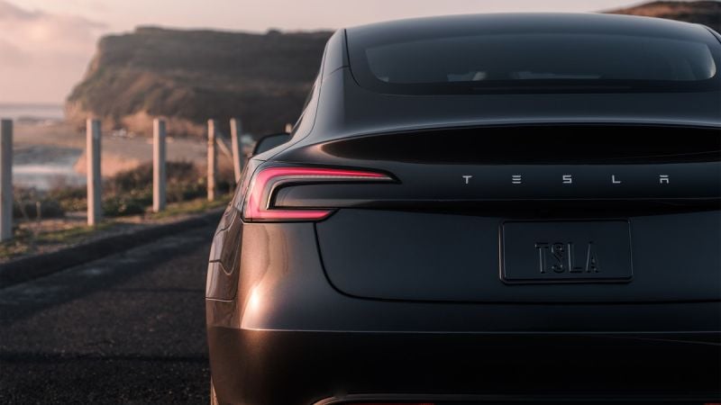 A sleek black Tesla Model 3 parked along a scenic coastline, with its logo and plate visible, under soft sunset lighting.