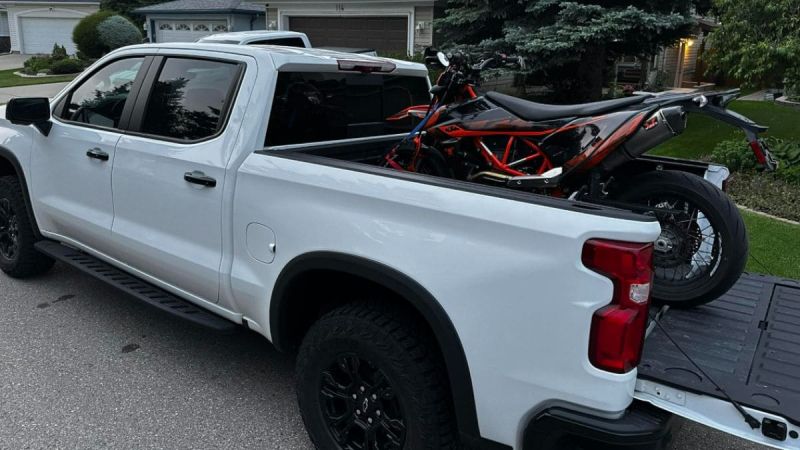 White Chevrolet Silverado pickup truck with black wheels transporting a red and black KTM motorcycle in its bed, parked in suburban driveway