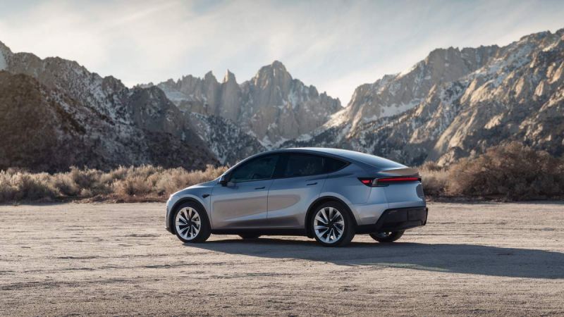 A silver 2026 Tesla Model Y Juniper parked on rocky terrain, with majestic snow-capped mountains in the background, under a clear blue sky.