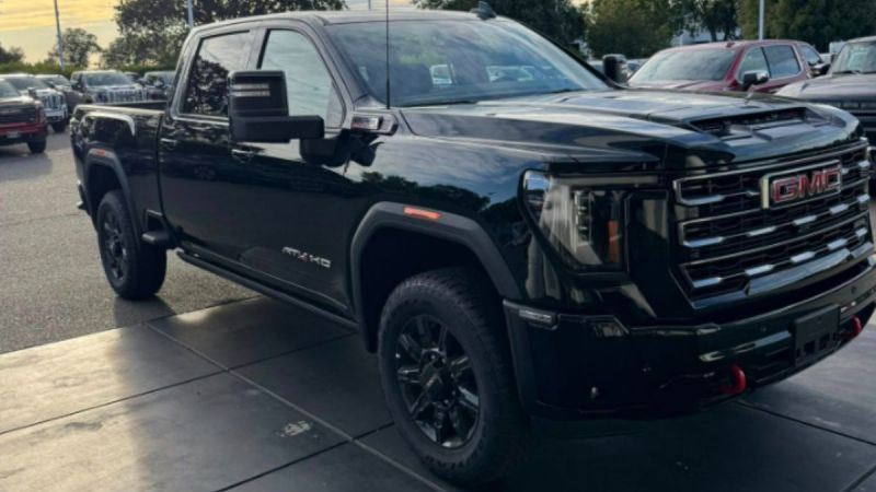A sleek black GMC Sierra 2500 H truck is parked under an overcast sky, showcasing its bold grille and robust stance. Surrounding trees and cars set a serene backdrop.