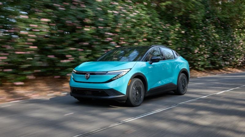 2026 Nissan LEAF electric vehicle driving on a tree-lined road, front three-quarter motion view.