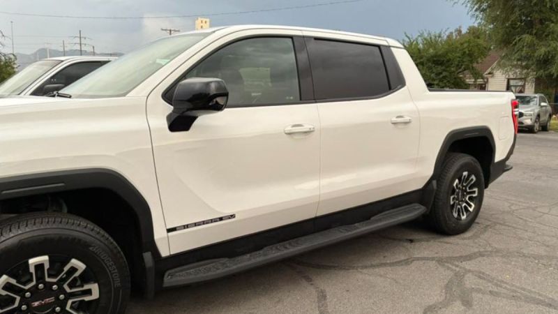 A white GMC Sierra EV truck parked, featuring sleek curves, dark rims, and tinted windows under a cloudy sky