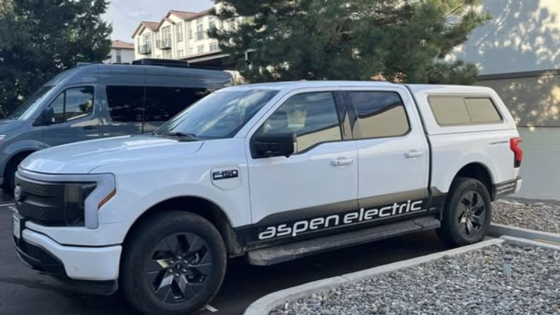 White Ford F-150 Lightning with "Aspen Electric" branding parked beside a van in a lot. Trees and residential buildings are visible in the background.