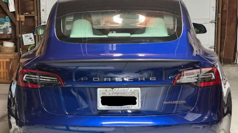 Blue Tesla Model 3 with a Porsche badge instead of the Tesla logo. Car is seen from the rear in a garage, the Dual Motor label is visible, reflecting a playful tone.