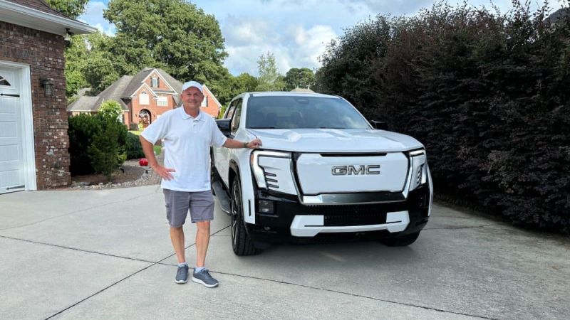 Perry is seen standing in a white polo shirt and cap smiling next to a white GMC Sierra EV in a suburban driveway. A brick house and greenery are visible in the background.