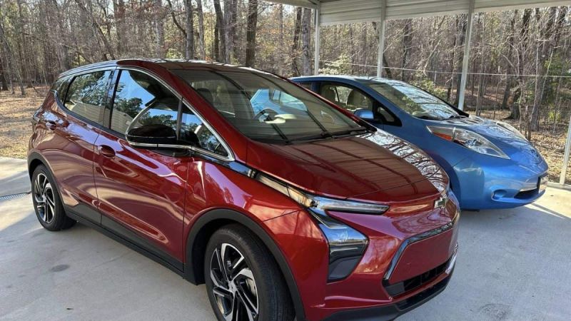 A maroon/red 2022 Chevrolet Bolt EUV is shown from a three-quarter front angle, parked next to a blue Nissan Leaf under a covered carport surrounded by trees.