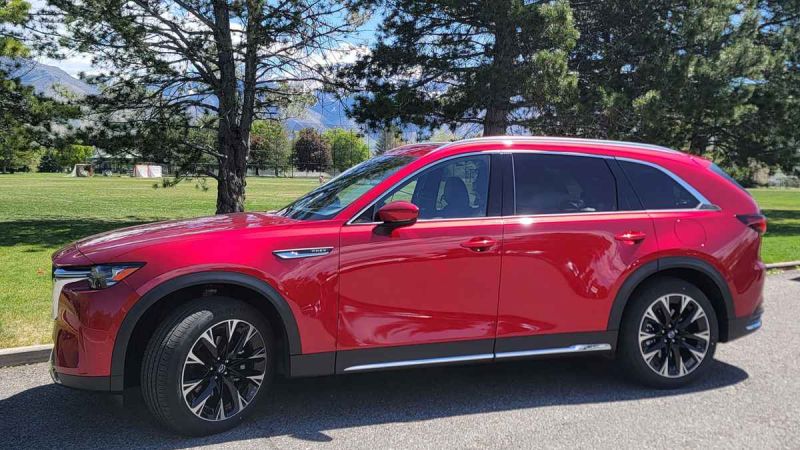 A Red 2025 Mazda CX-90 parked on a sunny day in a grassy area, with trees and mountains in the background.