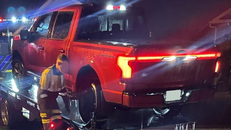 A red Ford truck is being loaded onto a tow truck at night, with rain falling and emergency lights flashing in the background.