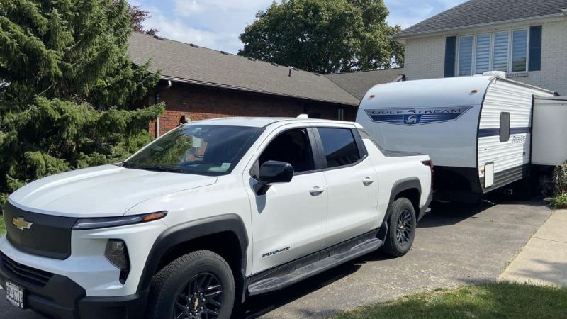 A white Chevrolet Silverado EV is parked in a driveway, hitched to a Gulf Stream travel trailer. A brick house and lush green trees are visible in the background.