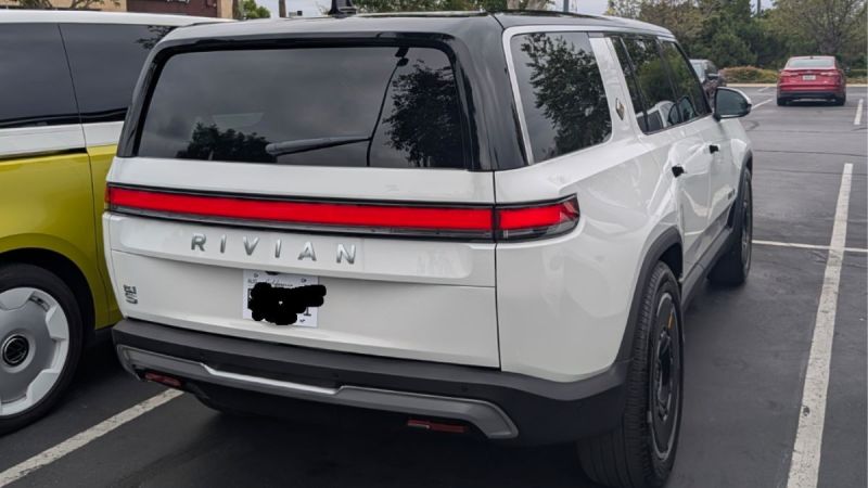 White 2025 Rivian R1S parked beside a yellow vehicle, showcasing its design and distinct rear light strip.
