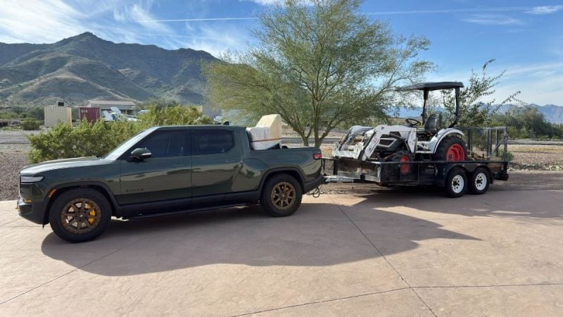 A black Rivian R1T electric pickup truck is shown from a side/front 3/4 angle, parked on a concrete pad with desert mountains visible in the background. The truck appears to be equipped with aftermarket bronze/gold wheels and a roof rack system.