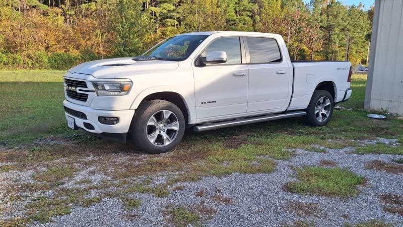 White 2024 Ram 1500 pickup truck shown from a front three-quarter view, parked on gravel with trees in the background, featuring chrome accents and alloy wheels.