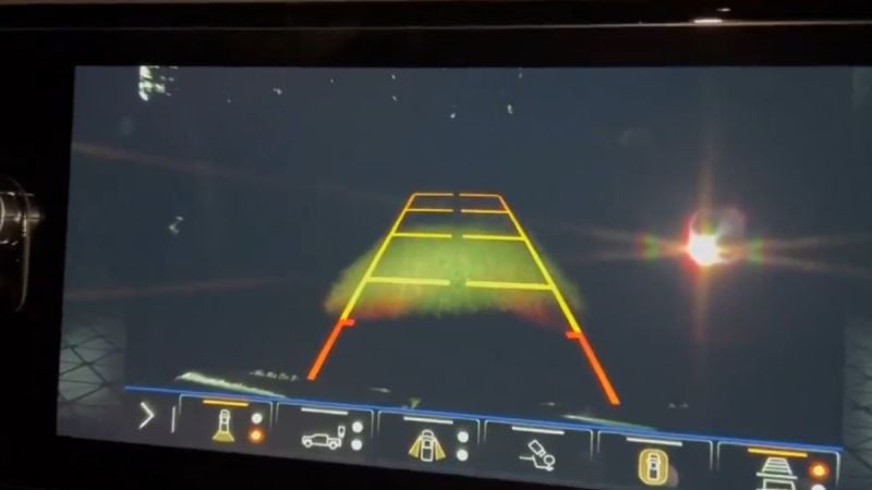 A car's rearview camera display shows a night scene with dark background and colorful guidelines for safe parking. A large lens flare is visible on the right.