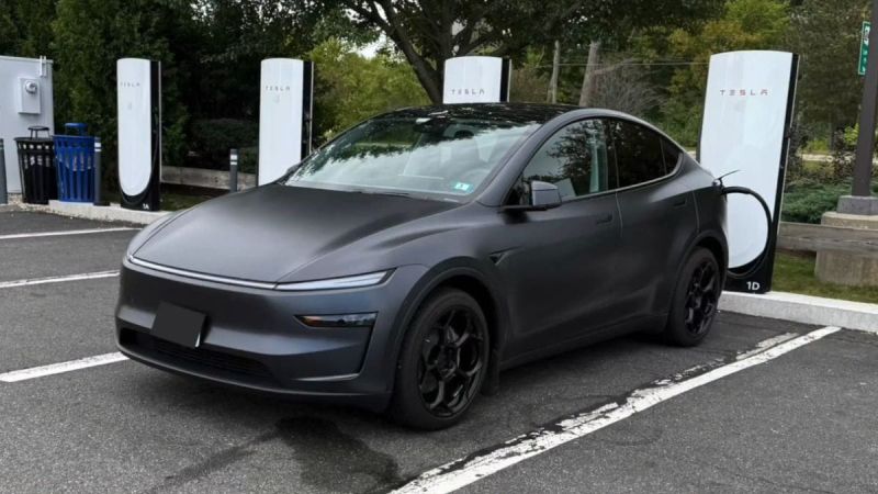 A matte black Tesla Model Y is parked at a Tesla charging station surrounded by greenery. The scene conveys a modern, eco-friendly vibe.