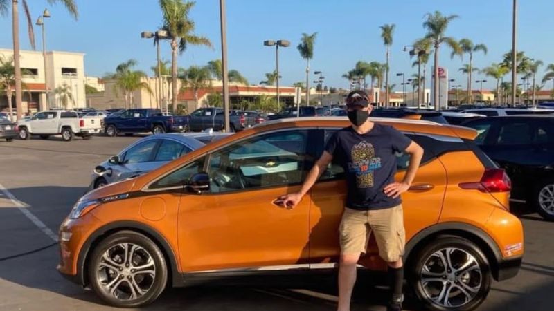 Scott wears a mask and is in casual clothing standing proudly beside an orange Chevy Bolt in a sunny parking lot filled with palm trees and parked vehicles.