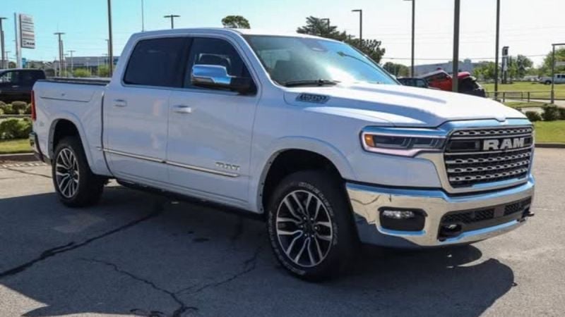 White Ram 1500 Limited parked outdoors, showcasing a shiny chrome grille and alloy wheels. The setting is a car lot with trees and blue sky in the background.