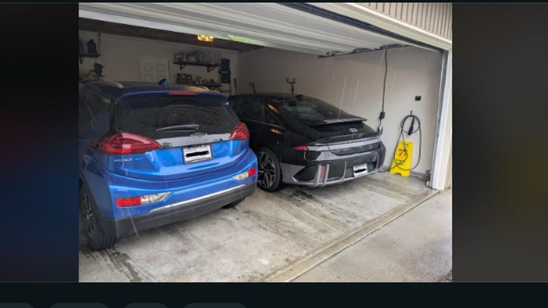 A blue Chevrolet Bolt EV and a black electric vehicle are parked in a garage, with cleaning supplies and shelves in the background.
